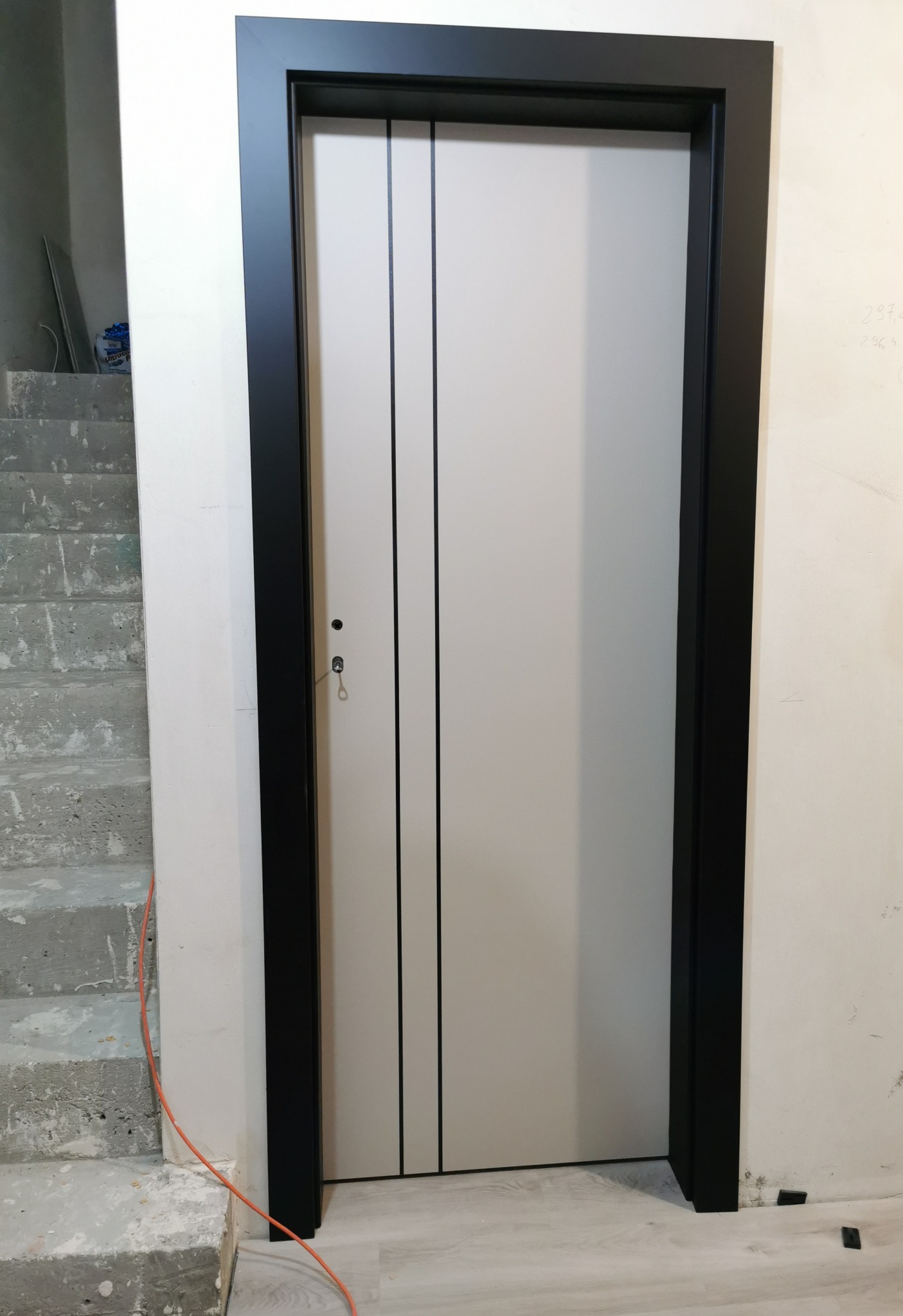 Zamknięte, białe drzwi wewnętrzne z czarną ościeżnicą, widoczne na tle betonowych schodów w surowym stanie.