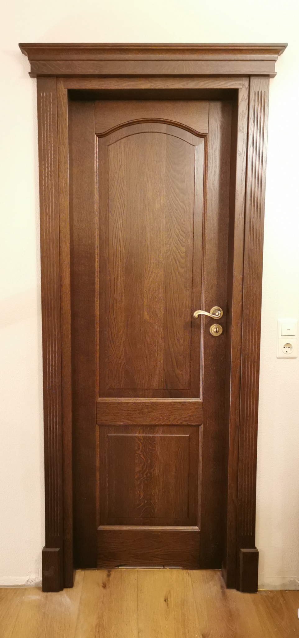 Ciemnobrązowe drzwi wewnętrzne z ozdobnymi listwami i mosiężną klamką, zamontowane w białej ścianie, widoczne gniazdko elektryczne.