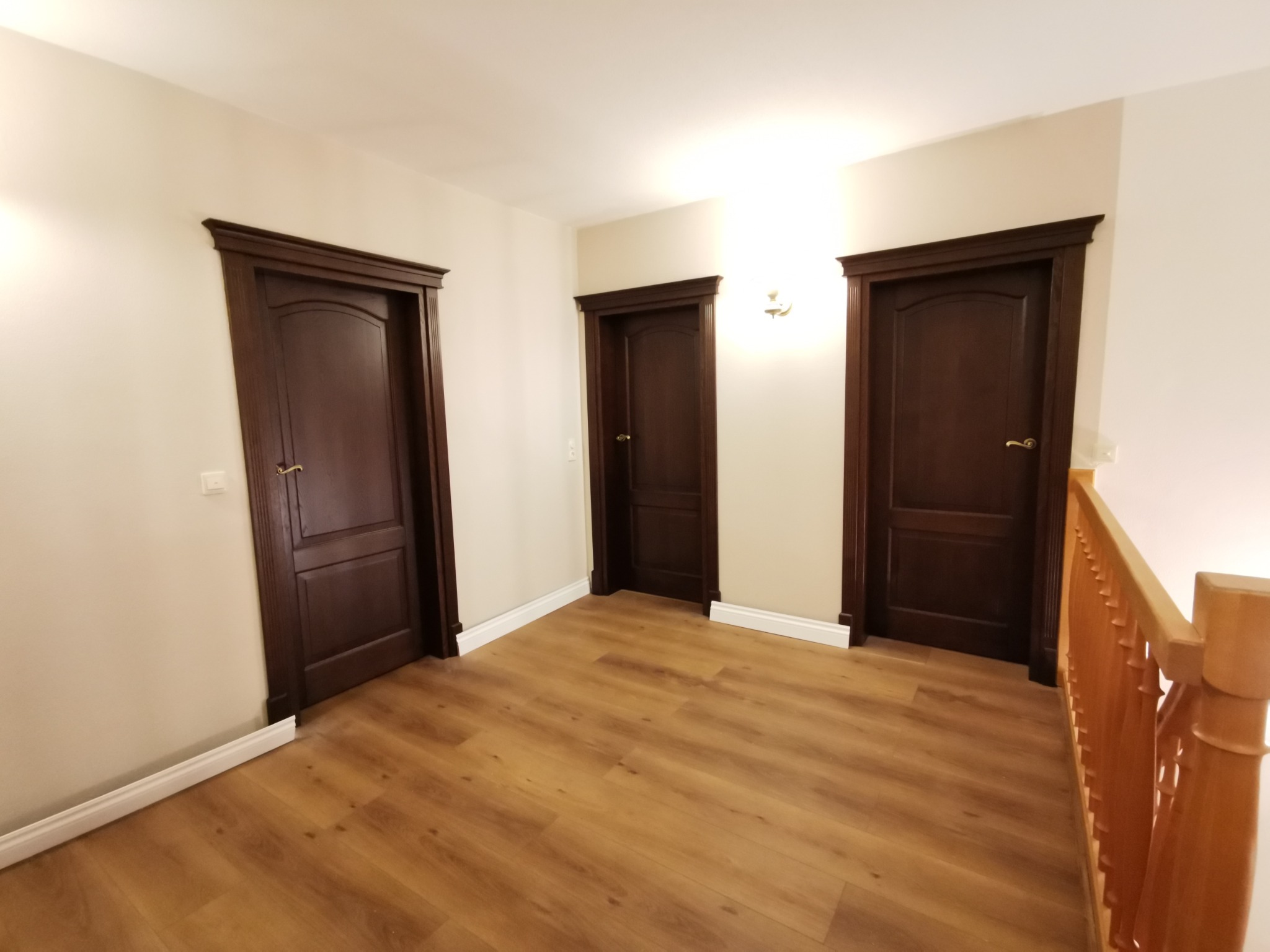 Korytarz z trzema ciemnobrązowymi drzwiami wewnętrznymi z dekoracyjnymi ościeżnicami, jasnymi ścianami i podłogą z paneli w odcieniach brązu, widoczna drewniana balustrada schodów.