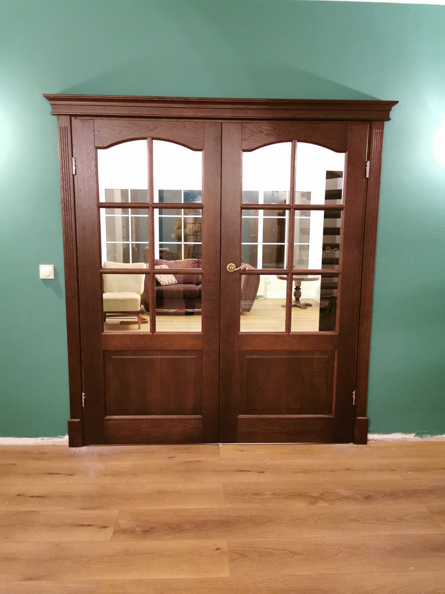 Dwuskrzydłowe, przeszklone drzwi wewnętrzne z ciemnego drewna, z widocznym fragmentem salonu w tle, zamontowane w pokoju o zielonych ścianach i jasnej podłodze.