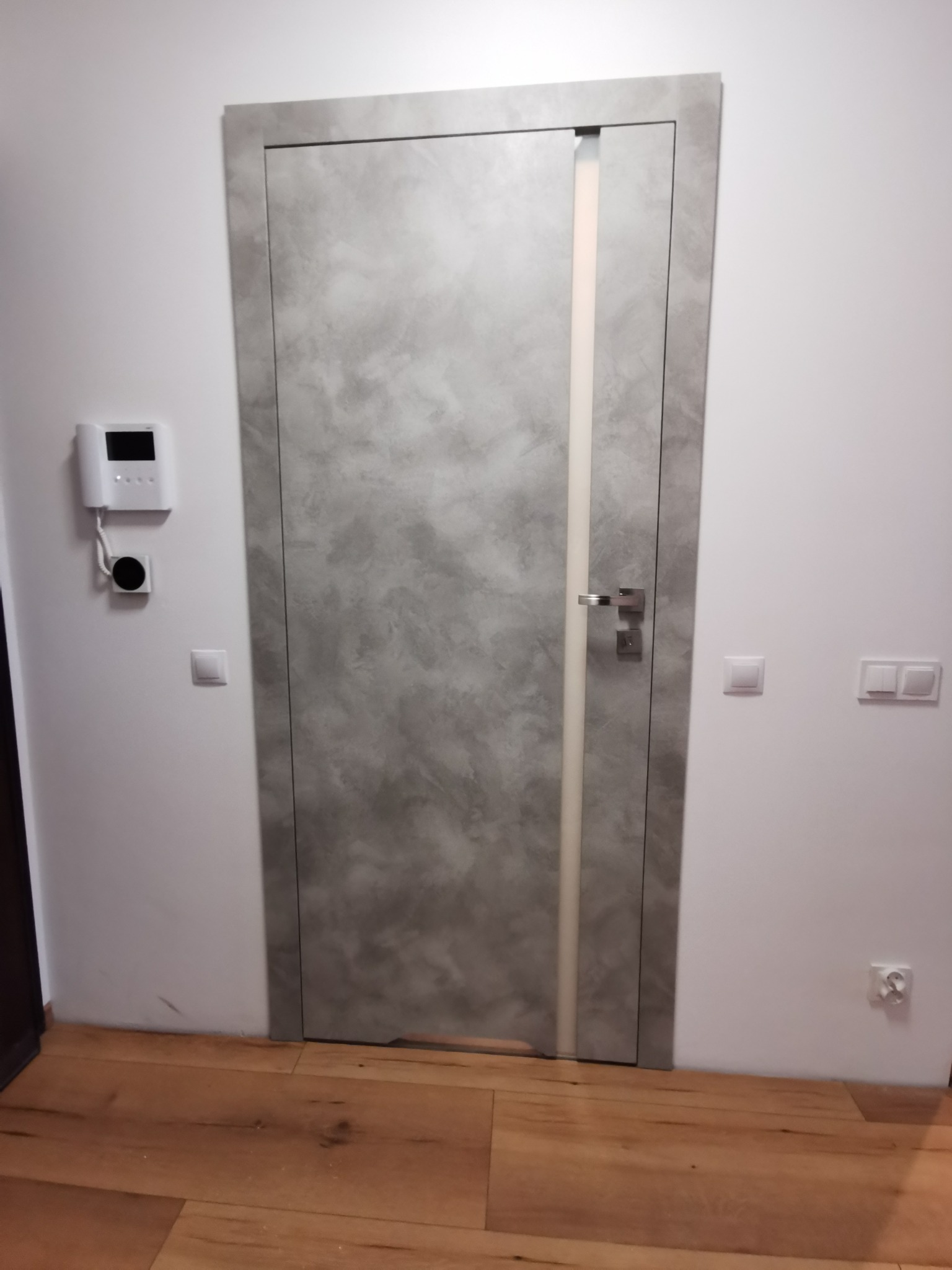 Szare drzwi wewnętrzne z pionowym, mlecznym przeszkleniem, zamontowane w białej ścianie z widocznym domofonem i włącznikami światła, na podłodze z paneli.