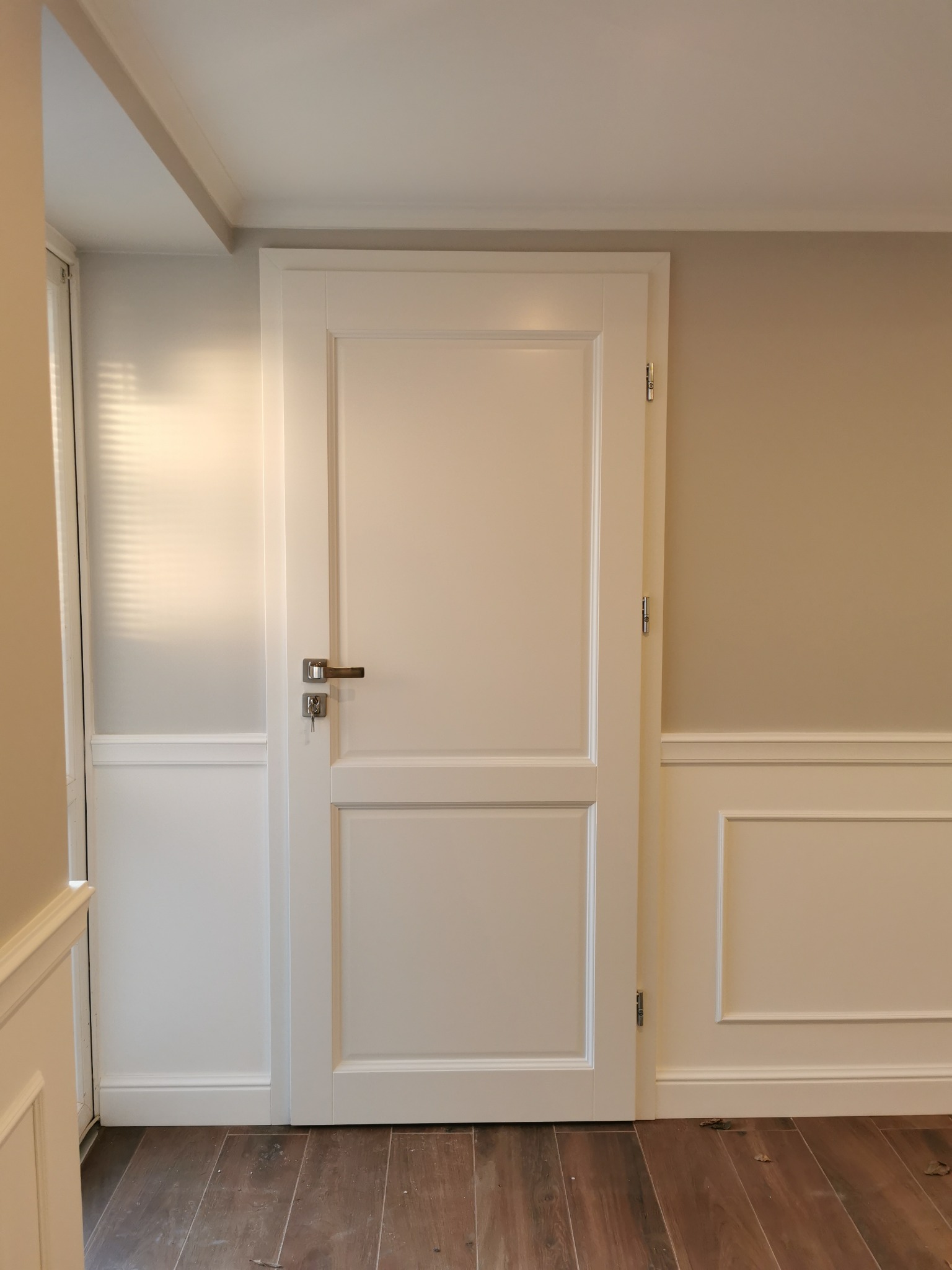 Białe drzwi wewnętrzne z prostokątnymi panelami i srebrną klamką, klucz w zamku, zamontowane w ścianie z dekoracyjną boazerią, widoczna podłoga z paneli.