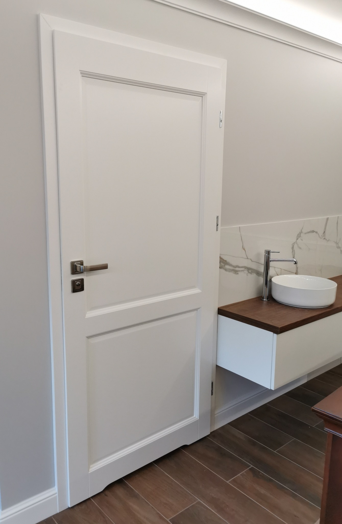 Białe drzwi wewnętrzne z metalową klamką w łazience z brązową podłogą i umywalką na blacie.