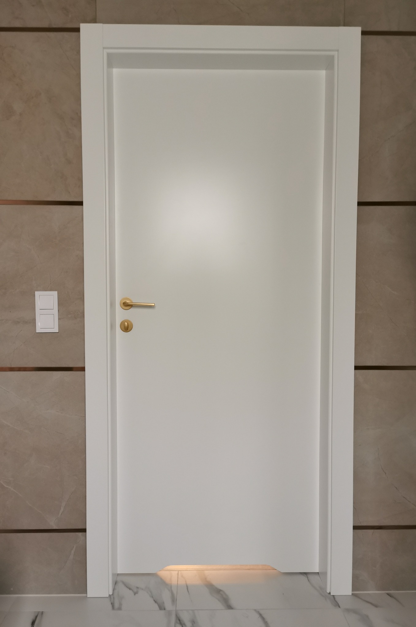 Białe drzwi wewnętrzne z złotą klamką na tle ściany z płytek imitujących marmur i marmurowej podłogi.