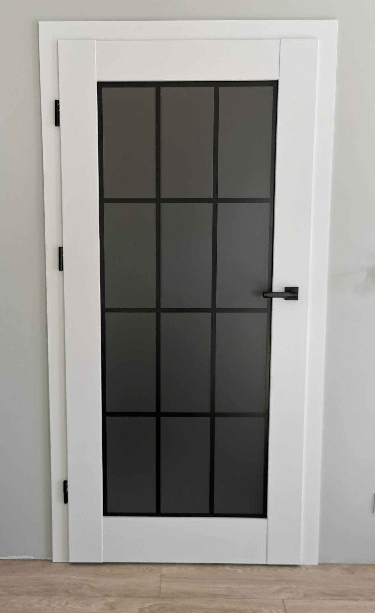 Białe drzwi wewnętrzne z czarnymi szprosami i klamką, na tle jasnoszarej ściany i drewnopodobnej podłogi.