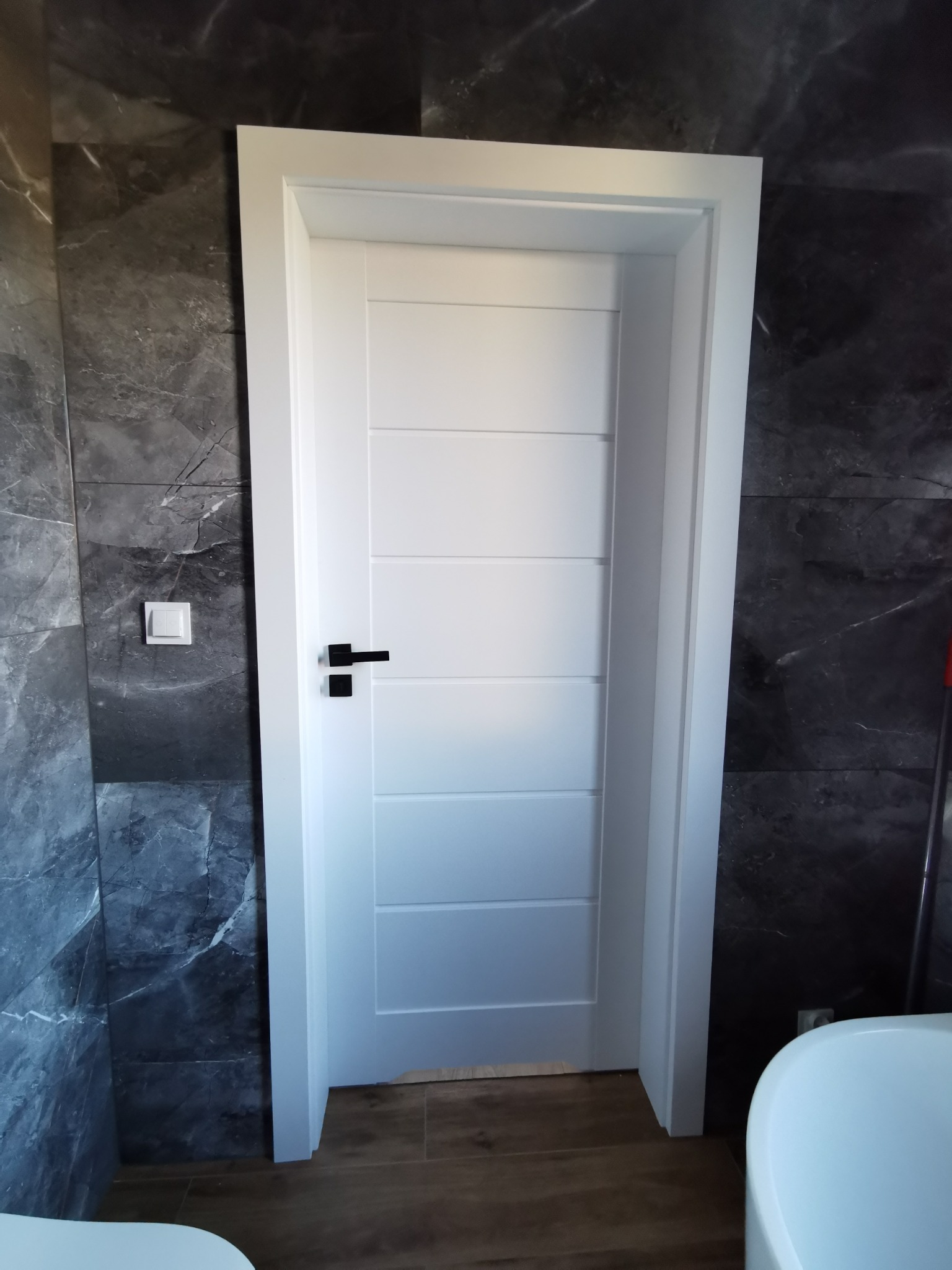 Białe drzwi wewnętrzne z czarną klamką w łazience z szarą marmurową okładziną ścienną i jasnym włącznikiem światła.