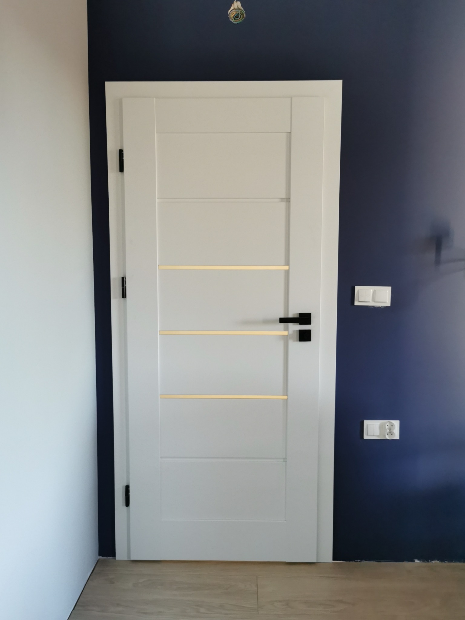 Białe drzwi wewnętrzne z czarną klamką i poziomymi, złotymi wstawkami, na tle ściany w kolorze granatowym i białym, widoczne gniazdka i włączniki.