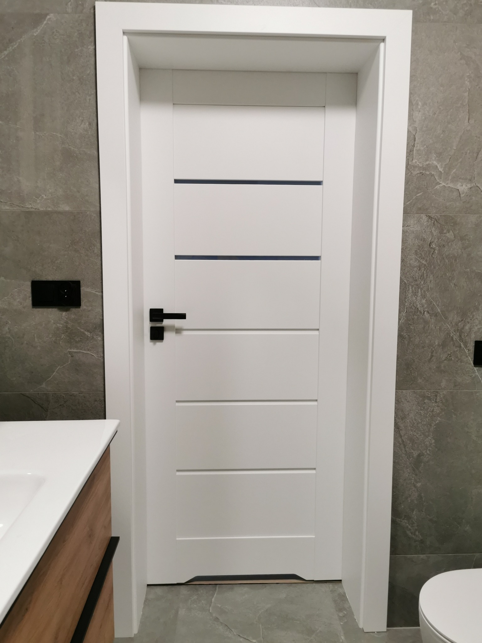 Białe drzwi wewnętrzne z czarną klamką i poziomymi, ciemnymi wstawkami na tle szarej ściany z imitacją kamienia, widoczny fragment umywalki.