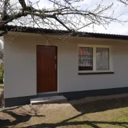 Otynkowana elewacja małego budynku gospodarczego z brązowymi drzwiami, oknem z firanką i ciemną podmurówką, widoczne drzewo z nagimi gałęziami i trawnik.