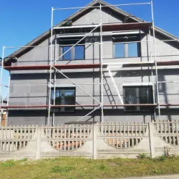 Dom jednorodzinny w trakcie ocieplania ścian zewnętrznych, widoczne szare płyty izolacyjne i rusztowanie, w tle błękitne niebo.