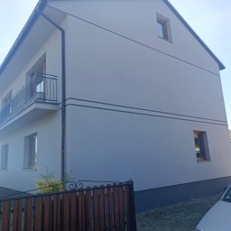 MOBUD Michał Owczarek - Dwukondygnacyjny dom z balkonem, po świeżym malowaniu elewacji na szaro, z ciemnymi pasami dekoracyjnymi. Widoczny fragment ogrodzenia i samochodu.