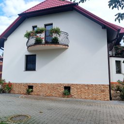 MOBUD Michał Owczarek - Biała elewacja domu z czerwonym dachem, balkonem z kwiatami i ceglaną podmurówką. Widoczne okna i roślinność. Ujęcie z poziomu chodnika.