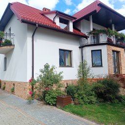 MOBUD Michał Owczarek - Biały dom z czerwoną dachówką i ceglaną podmurówką, balkony z kwiatami, widok z zewnątrz, kostka brukowa na podjeździe, roślinność w ogrodzie.