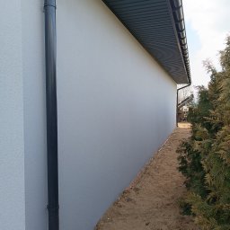 MOBUD Michał Owczarek - Świeżo otynkowana, jasnoszara ściana budynku z zamontowaną rynną spustową. Widoczny fragment dachu i roślinność w tle. Tekstura tynku barankowego.