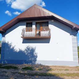 MOBUD Michał Owczarek - Jasnoszara elewacja domu z brązowym dachem i balkonem z kutą balustradą, na tle błękitnego nieba z chmurami. Widoczne detale wykończenia i świeżo pomalowana fasada.