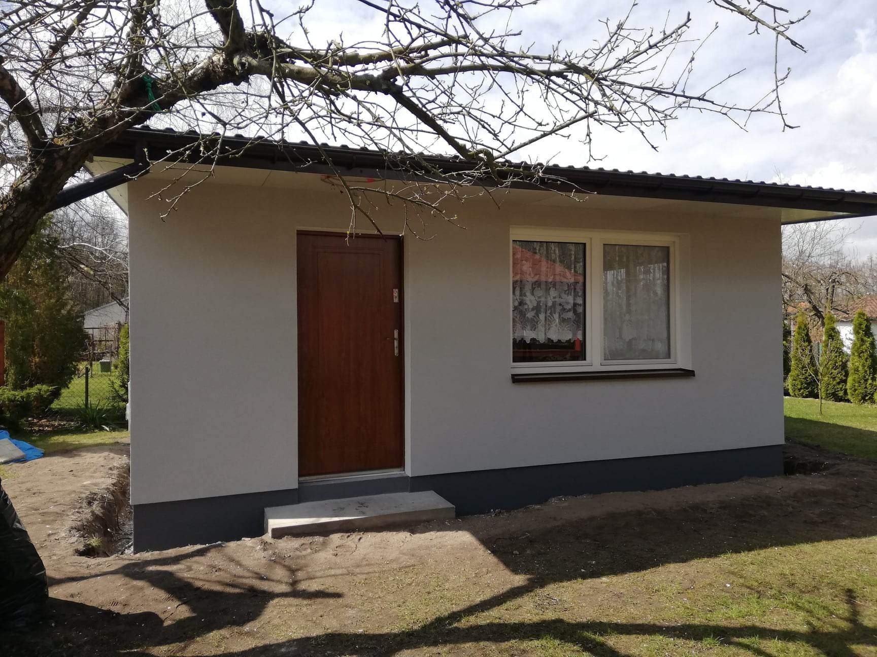 Otynkowana elewacja małego budynku gospodarczego z brązowymi drzwiami, oknem z firanką i ciemną podmurówką, widoczne drzewo z nagimi gałęziami i trawnik.