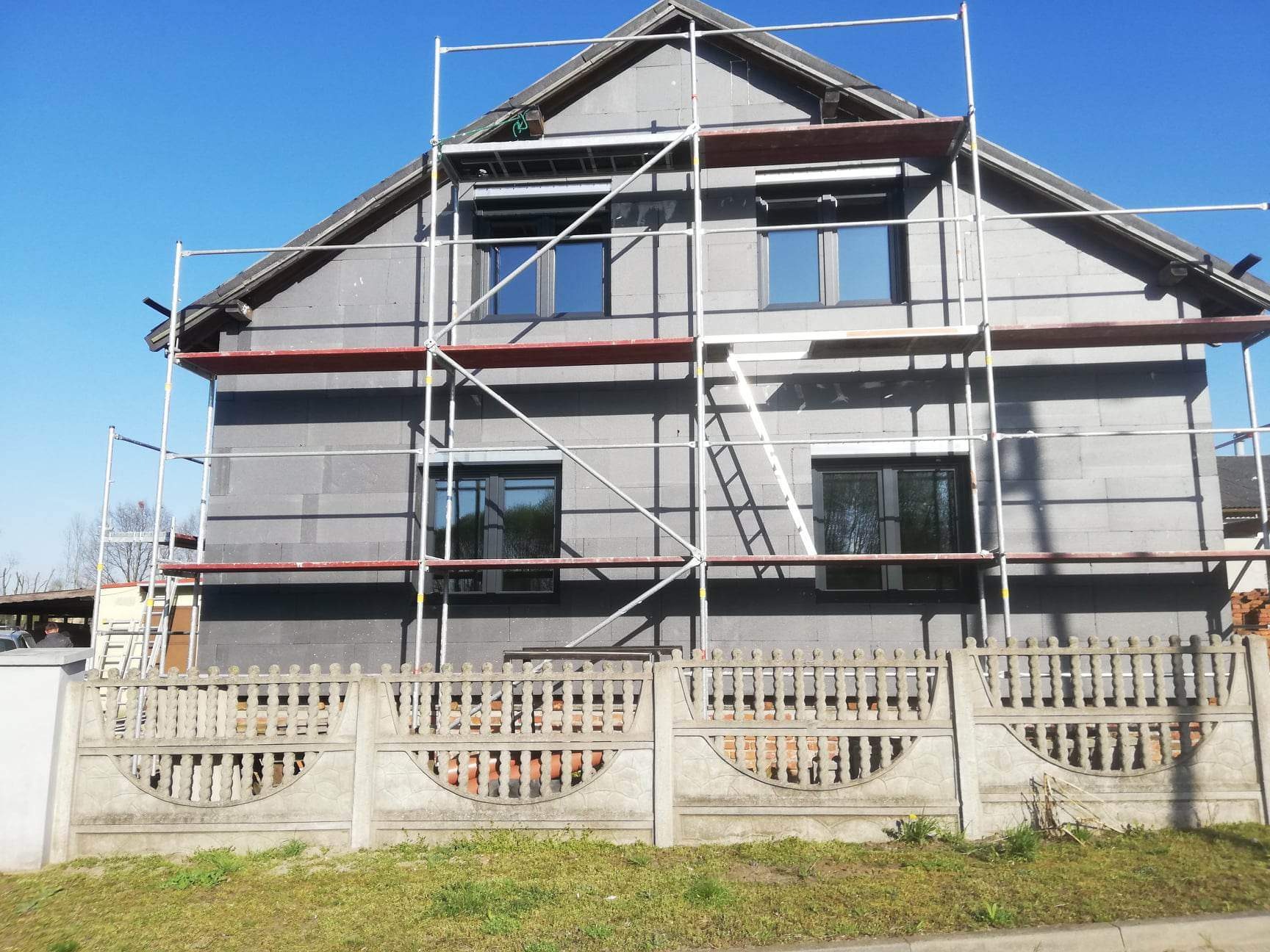 Dom jednorodzinny w trakcie ocieplania ścian zewnętrznych, widoczne szare płyty izolacyjne i rusztowanie, w tle błękitne niebo.