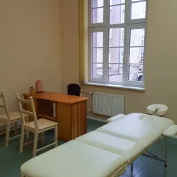 Jasne wnętrze gabinetu z białym łóżkiem do zabiegów, biurkiem z lampą solną i widokiem na budynek z okna.