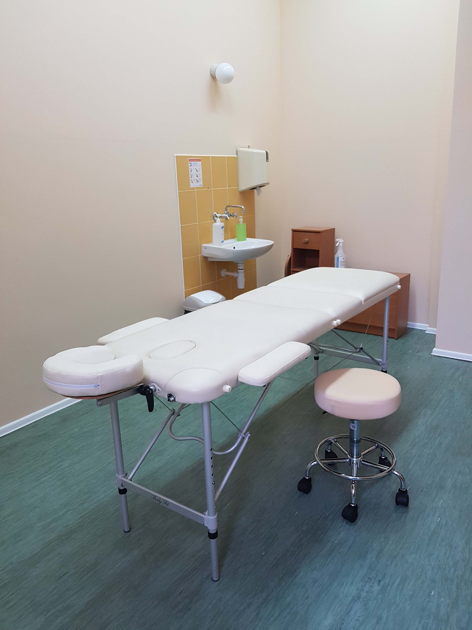 Jasne wnętrze gabinetu z profesjonalnym, białym stołem do zabiegów z regulowanym zagłówkiem i podłokietnikami, obok taboret na kółkach, umywalka z dozownikiem mydła i szafka w tle.