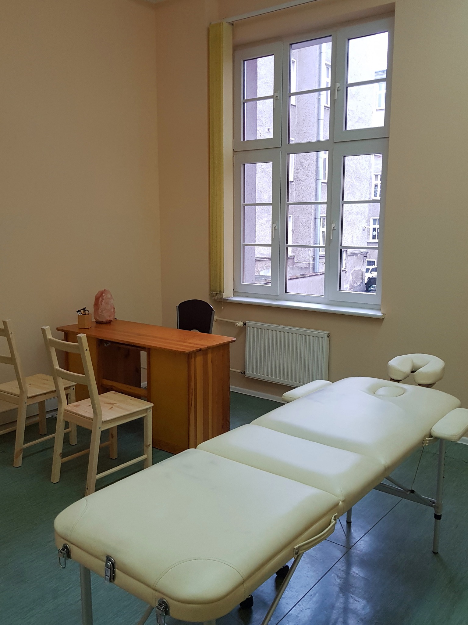 Jasne wnętrze gabinetu z białym łóżkiem do zabiegów, biurkiem z lampą solną i widokiem na budynek z okna.