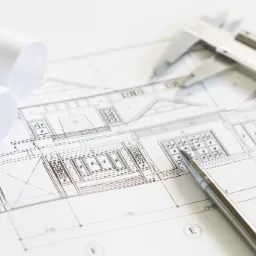 Schemat architektoniczny domu z naniesionymi wymiarami, leżący na białym tle, z dwoma zrolowanymi arkuszami papieru, suwmiarką i metalowym długopisem wskazującym na detal okna.