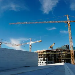 Widok na plac budowy z trzema żurawiami wieżowymi na tle błękitnego nieba, w tle powstające budynki z widoczną konstrukcją szkieletową i elewacją w trakcie montażu.