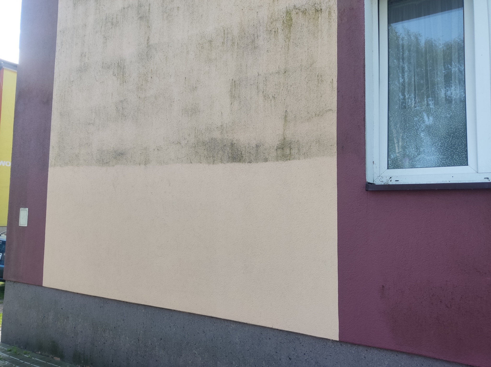 Fragment elewacji budynku z widoczną różnicą w czystości powierzchni, górna część ściany jest zabrudzona, dolna świeżo umyta, obok okno z zaparowaną szybą i ściana pomalowana na kolor bordowy.