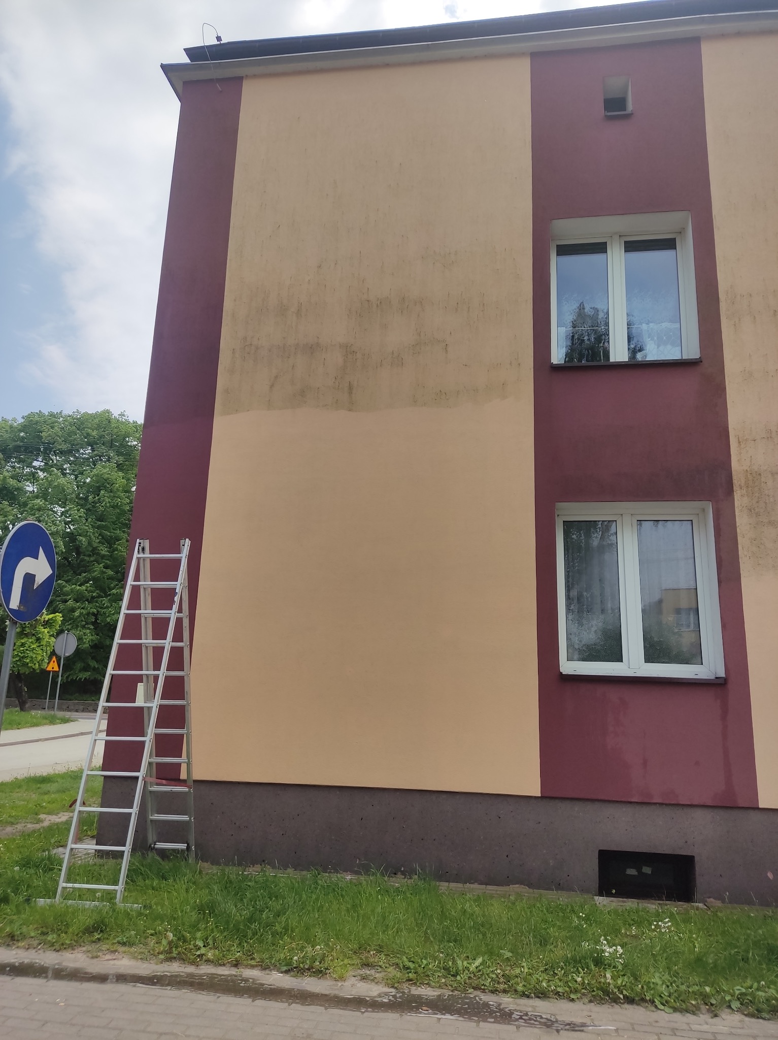 Dwukolorowa elewacja budynku w trakcie odświeżania, z widoczną różnicą w kolorze między umytą a brudną powierzchnią, drabina oparta o ścianę, znak drogowy w tle.