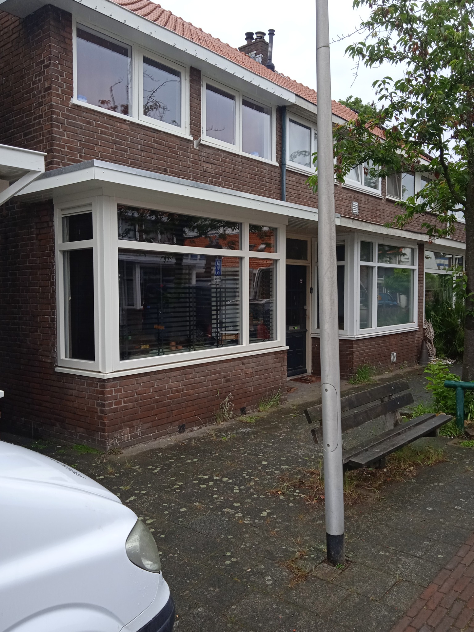 Brązowy dom z cegły z białymi ramami okiennymi, widok z zewnątrz, częściowo zasłonięty przez zaparkowany biały samochód i słup oświetleniowy. Na pierwszym planie widoczna drewniana ławka.