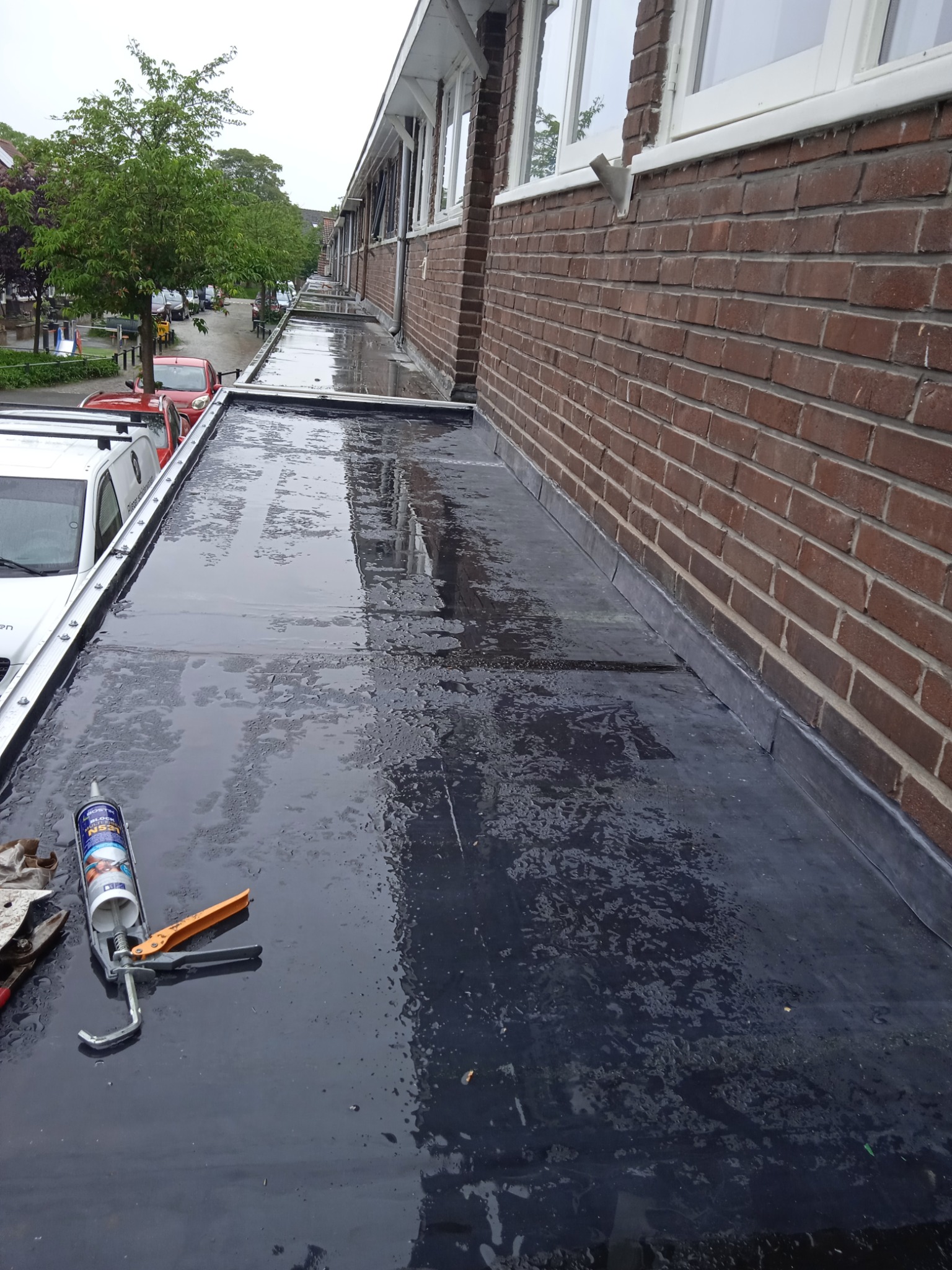 Widok z góry na płaski, mokry dach pokryty papą, z narzędziami do uszczelniania (pistolet do silikonu i tuba z uszczelniaczem) leżącymi w rogu, obok ściany budynku z cegły i okien.