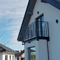 Nowoczesny dom z balkonem zabezpieczonym prostą, czarną metalową konstrukcją, widok z dołu na tle błękitnego nieba.