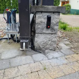 Szczegółowe ujęcie zamontowanego napędu bramy przesuwnej z widocznymi przewodami, czujnikiem zbliżeniowym na kamiennym słupku, oraz fragmentem bramy, kostką brukową i elementami ogrodu w tle.