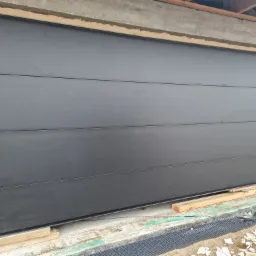 Zamknięta, segmentowa brama garażowa w kolorze antracytowym, widoczna od strony zewnętrznej, w trakcie montażu w surowym otworze, z widocznymi deskami podtrzymującymi.