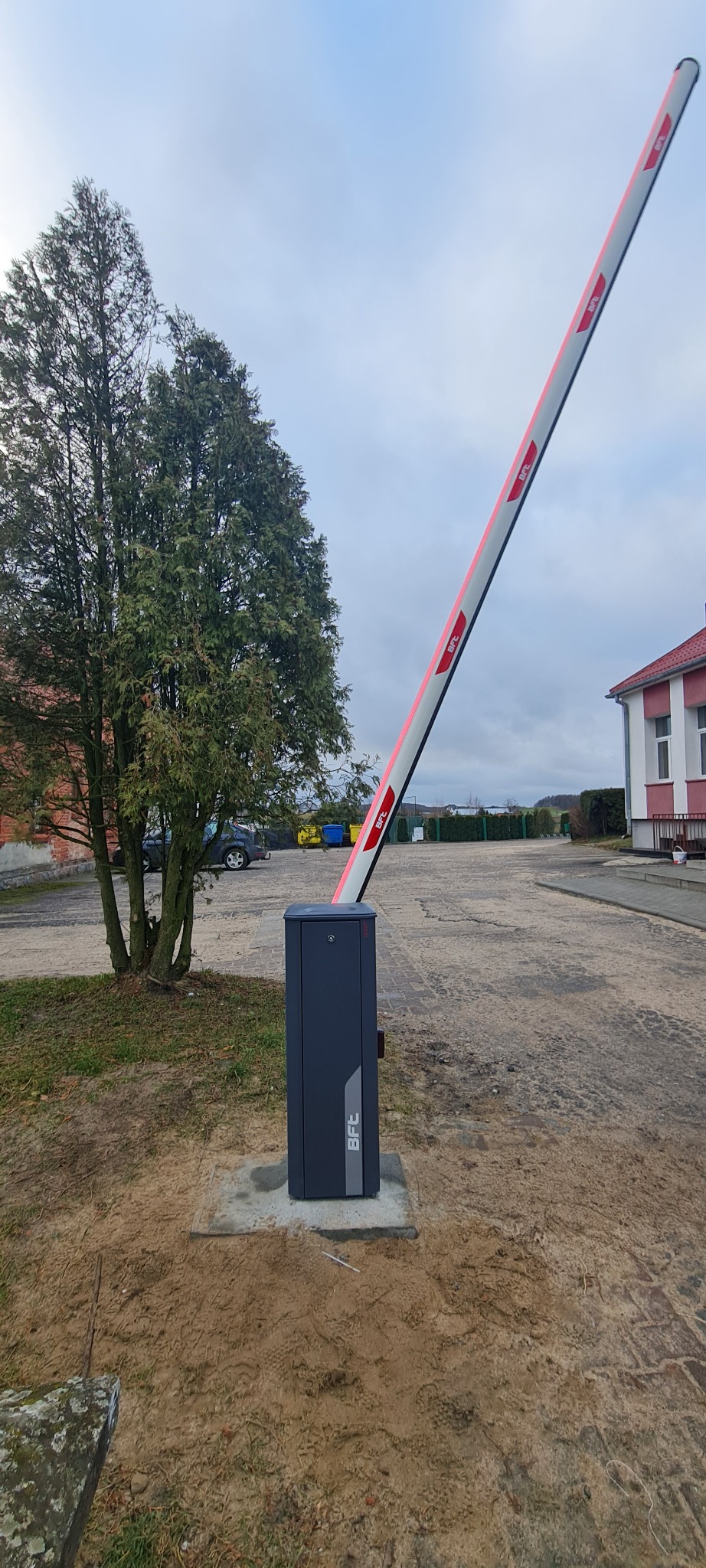 Podniesiona szlabanowa zapora drogowa BFC z czerwonymi odblaskami, widoczna na tle parkingu i drzewa