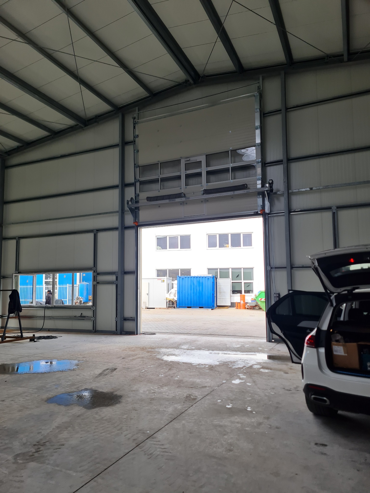 Wnętrze hali z otwartą automatyczną bramą segmentową, widoczny samochód z otwartym bagażnikiem i kałuże na podłodze.