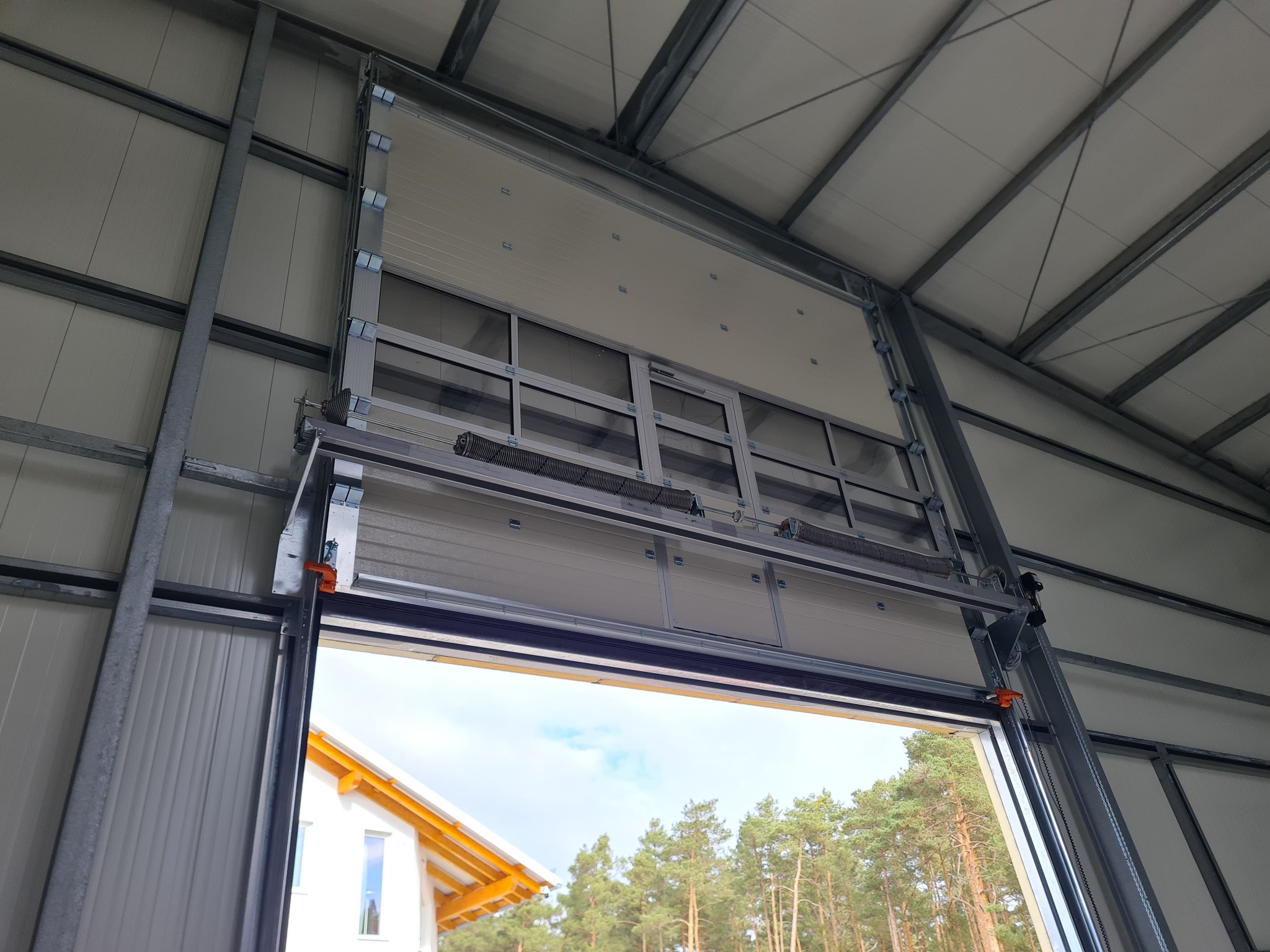 Segmentowa brama garażowa w trakcie otwierania, widok z wnętrza garażu na dom i drzewa na zewnątrz.