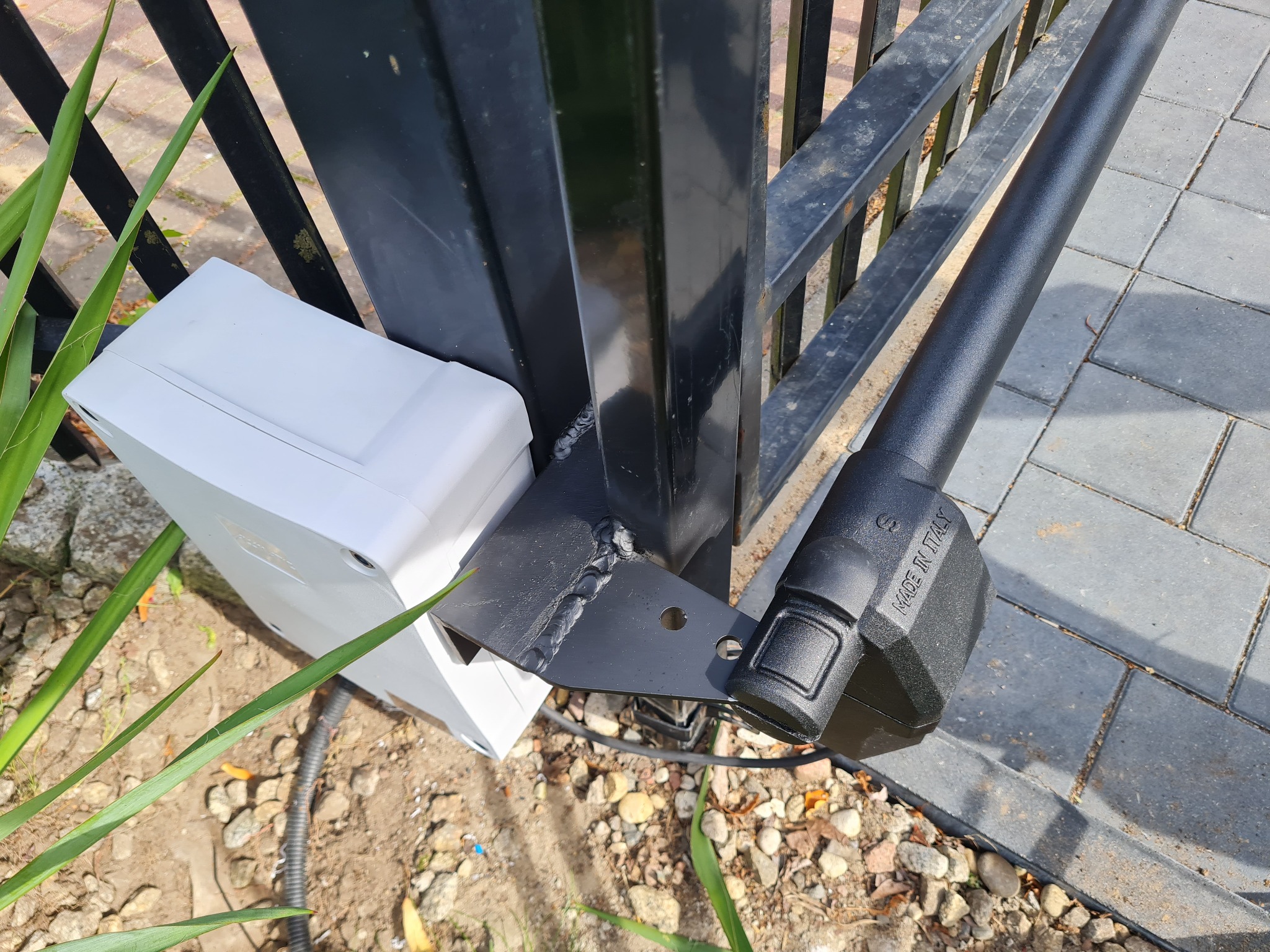 Zamknięcie bramy z napędem elektrycznym, widoczny siłownik z napisem 'Made in Italy' oraz biała obudowa sterownika, zamontowane na czarnej, metalowej konstrukcji bramy.