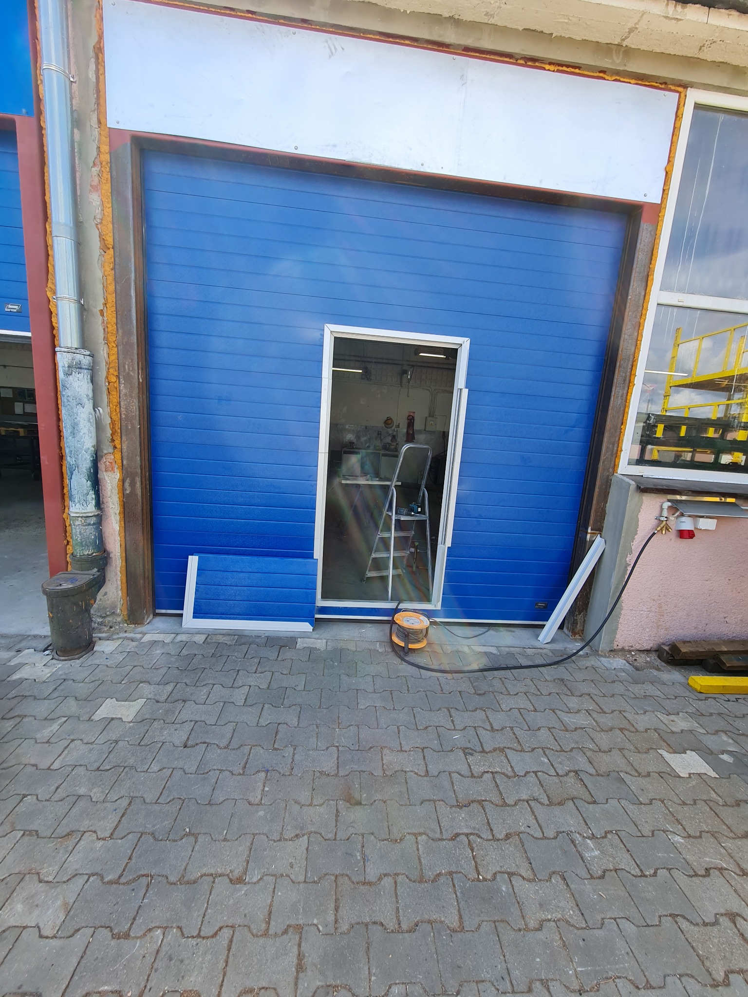 Niebieska segmentowa brama garażowa z częściowo zamontowanymi drzwiami wejściowymi, widoczna drabina wewnątrz garażu i zwinięty kabel zasilający na zewnątrz.