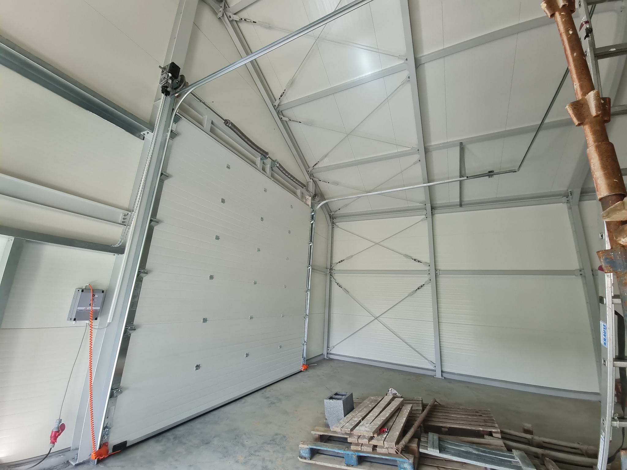 Wnętrze hali z częściowo otwartą białą bramą segmentową, widoczne elementy konstrukcyjne i napęd bramy, na podłodze paleta z deskami i betonowy bloczek.