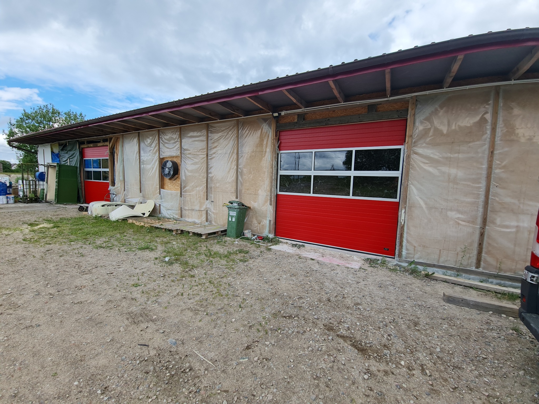 Czerwona segmentowa brama garażowa z oknami, zamontowana w budynku z elewacją w trakcie wykończenia, widoczne rusztowania i folie ochronne.