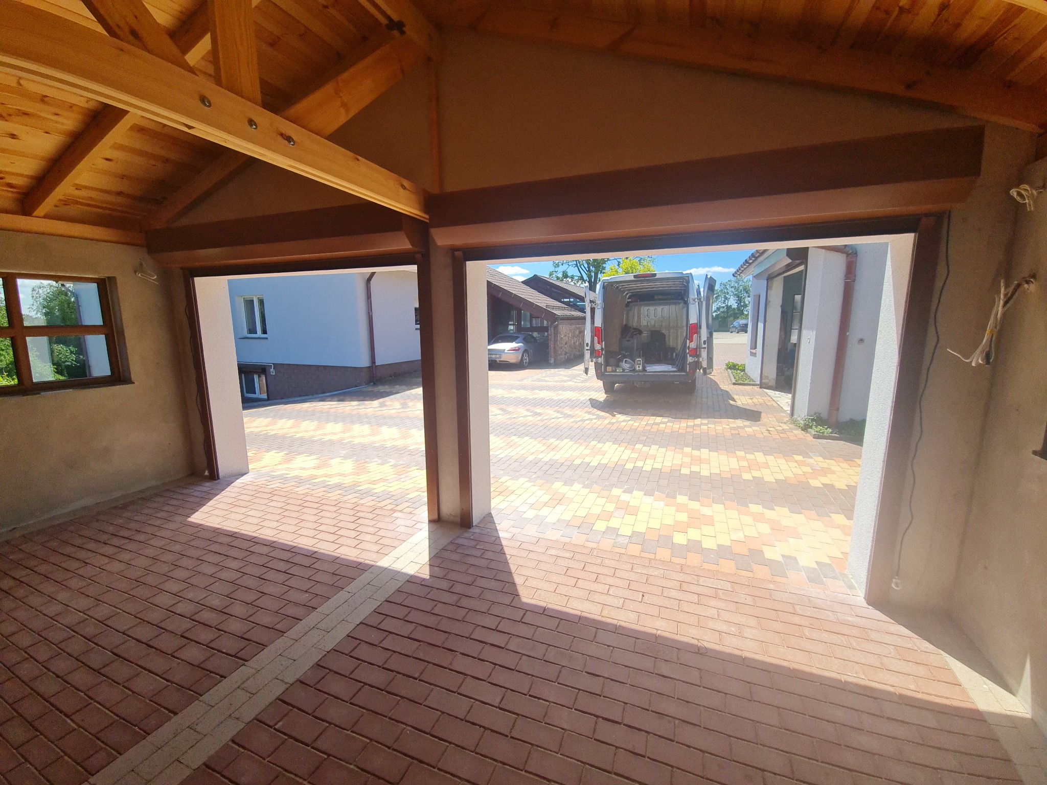Widok z wnętrza garażu z drewnianym stropem na podjazd z kostki brukowej i częściowo otwarte bramy rolowane; w tle zaparkowany biały bus.
