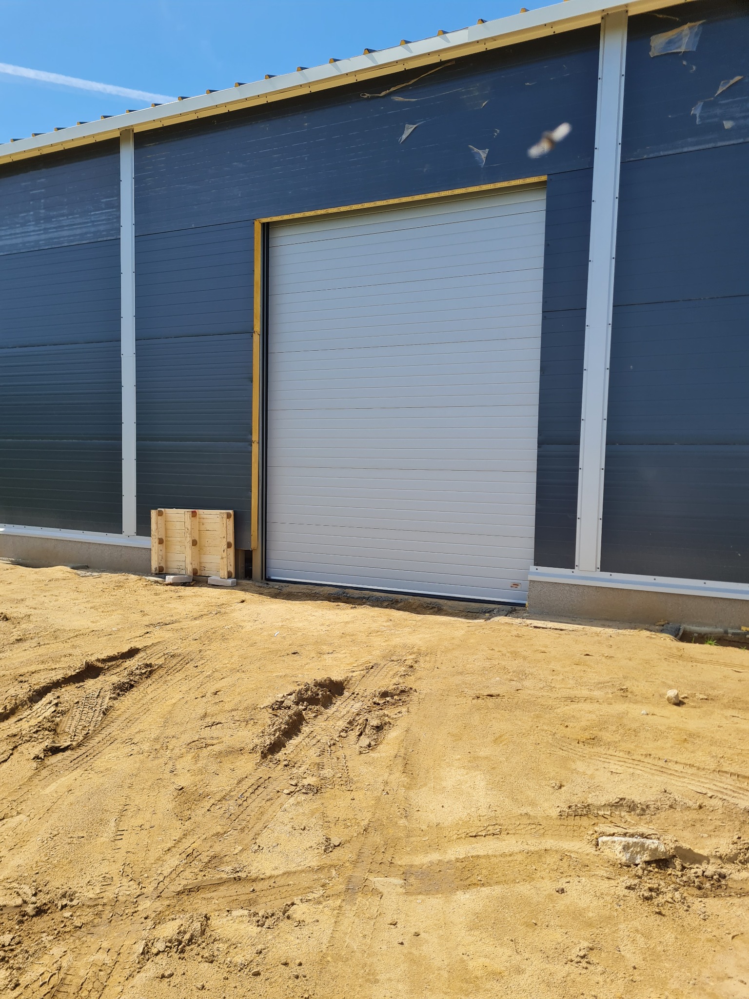 Zamknięta, segmentowa brama przemysłowa w hali o szarych ścianach, widoczne ślady opon na piaszczystym podłożu i drewniane elementy zabezpieczające.