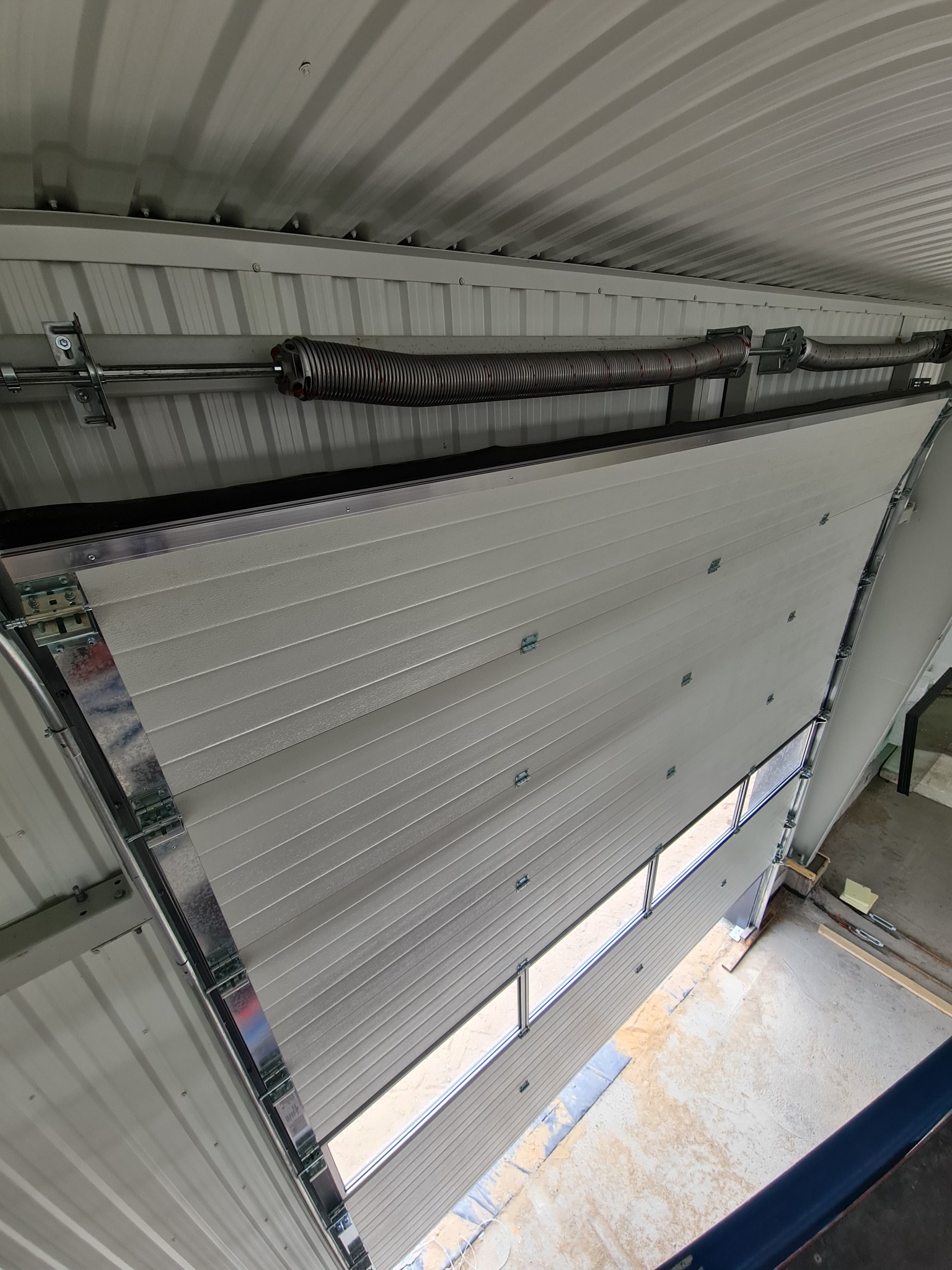 Otwarta segmentowa brama garażowa z widocznym mechanizmem sprężynowym i panelem z oknami, sfotografowana z góry.