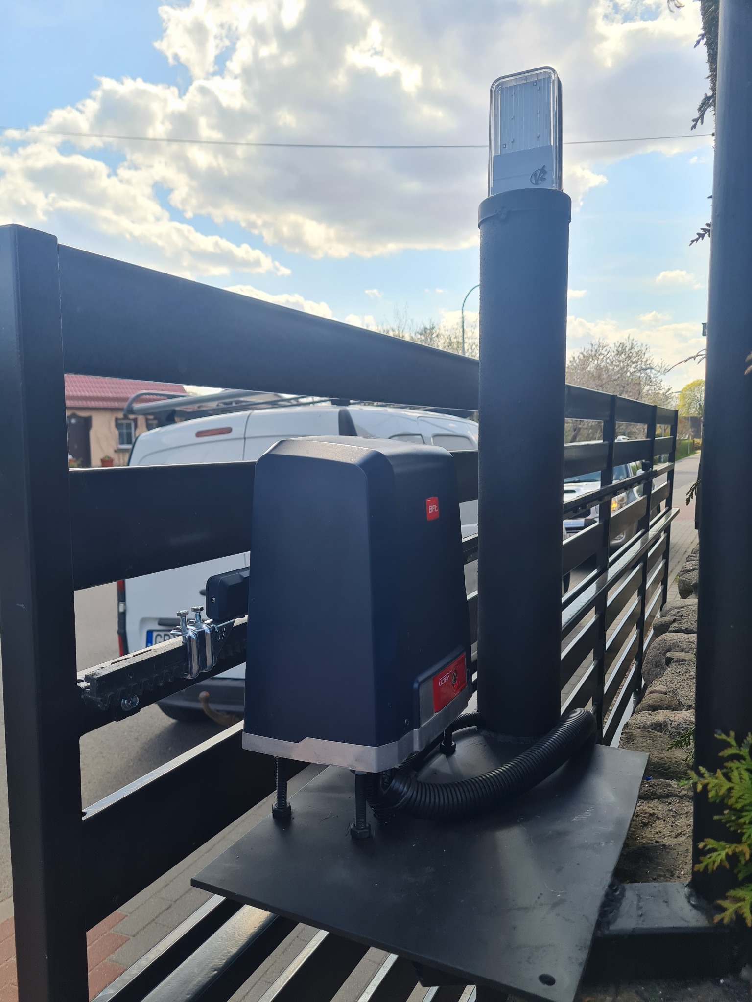 Zamontowany napęd do bramy skrzydłowej BFT Ultrix na tle błękitnego nieba z obłokami, widoczny także słupek z lampą ostrzegawczą.