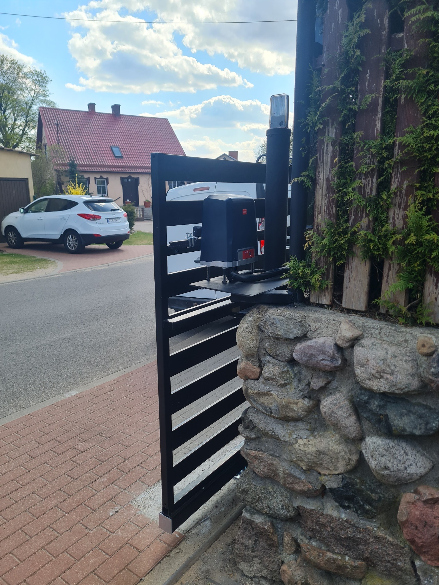 Czarna, pozioma brama przesuwna z napędem, zamontowana na kamiennym murku; w tle dom z czerwoną dachówką i biały samochód.
