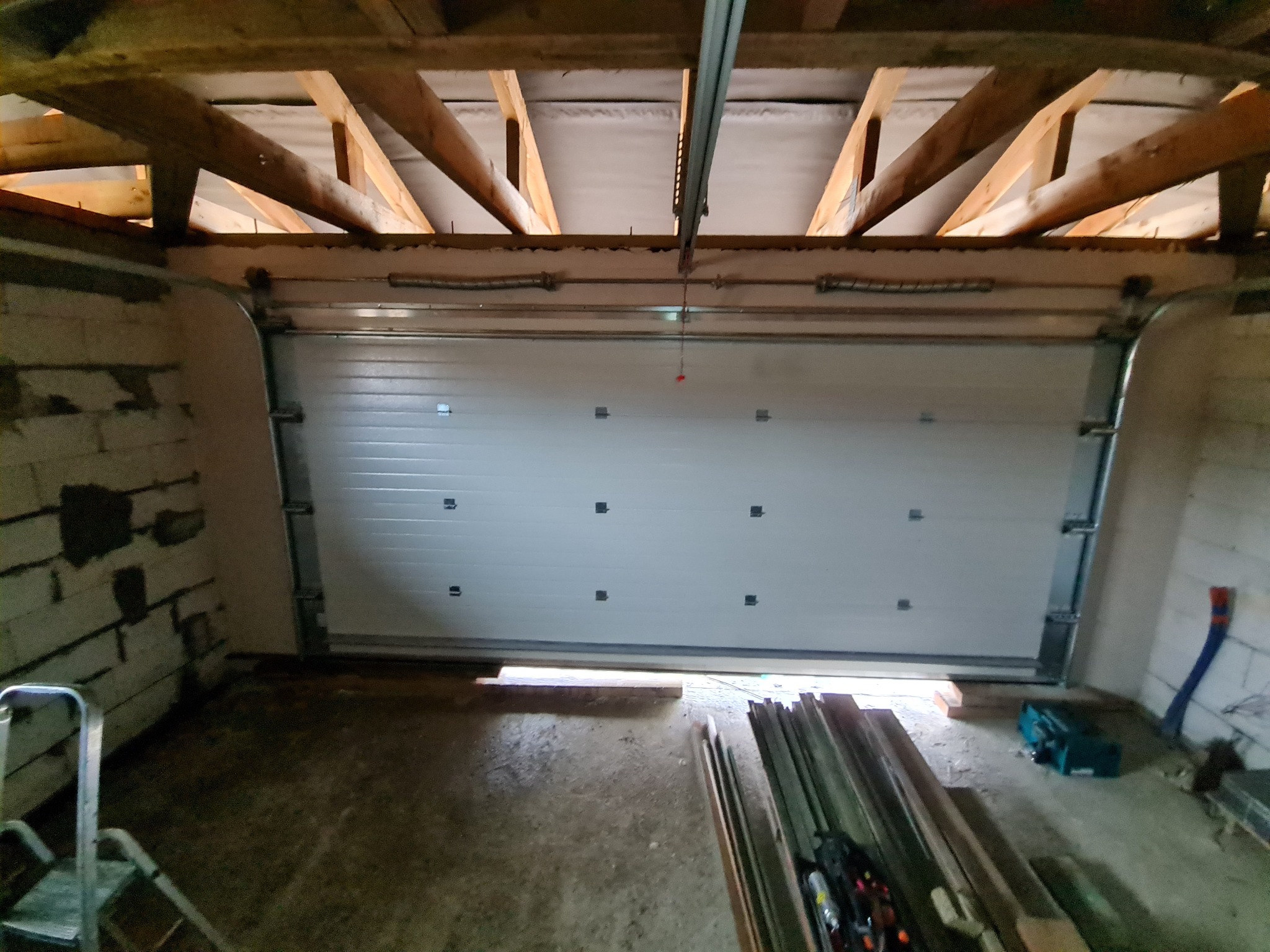 Montaż segmentowej bramy garażowej z widoczną konstrukcją drewnianą dachu i narzędziami na podłodze.