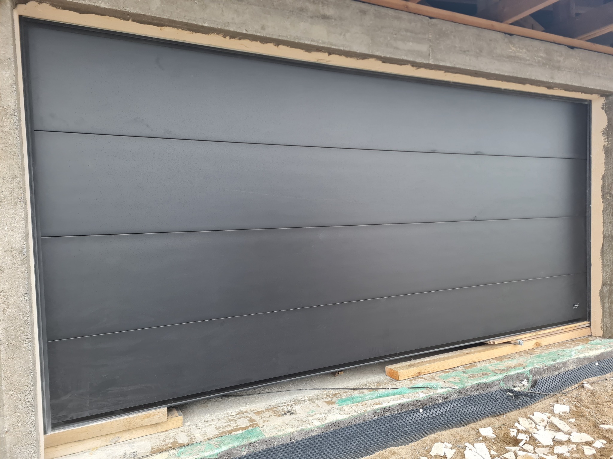 Zamknięta, segmentowa brama garażowa w kolorze antracytowym, widoczna od strony zewnętrznej, w trakcie montażu w surowym otworze, z widocznymi deskami podtrzymującymi.