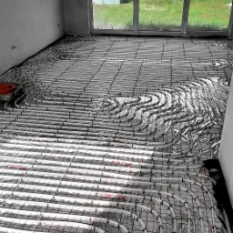 Białe rury ogrzewania podłogowego ułożone na siatce z folią odblaskową w surowym pomieszczeniu z dużym oknem na tle zieleni.