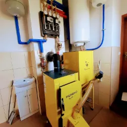 Żółty piec na pellet Pereko Q-Pell z podajnikiem OmPell, podłączony do instalacji grzewczej z zbiornikiem wyrównawczym i zasobnikiem ciepłej wody, w kotłowni wyłożonej kafelkami.