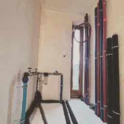 Instalacja wodno-kanalizacyjna i grzewcza w trakcie montażu, widoczne rury w izolacji na podłodze i piony rur przy ścianie, filtr wody z manometrami i zaworami.